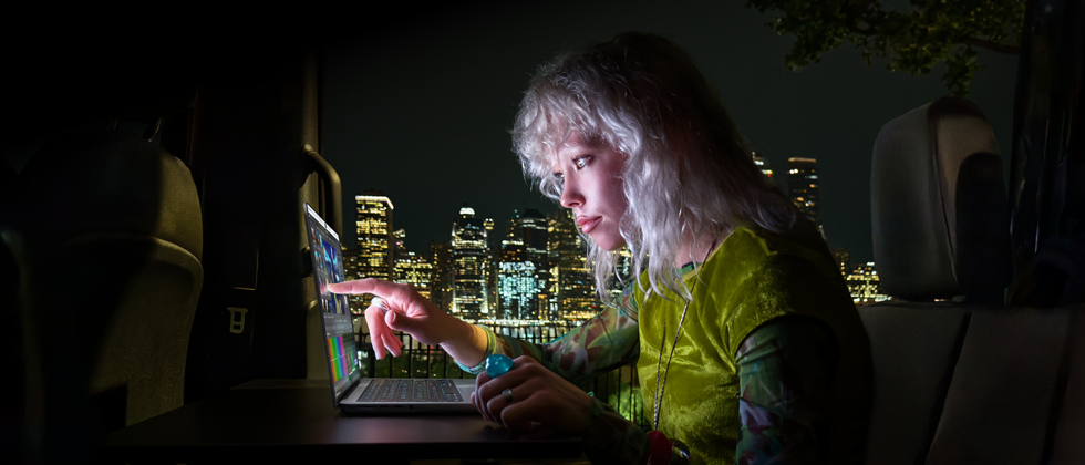 Eine Person sitzt auf dem Rücksitz eines Vans, durch die geöffnete Beifahrertür ist die Skyline einer Stadt bei Nacht zu sehen, ein MacBook Pro mit PROLED XDR Display auf einem Schreibtisch, die Person wird vom Bildschirm beleuchtet, während sie das Touch Feature nutzt, um ein Video in DaVinci Resolve Studio zu bearbeiten