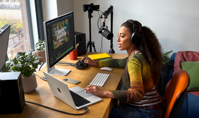 Eine Frau arbeitet bei sich zu Hause an ihrem Schreibtisch mit einem MacBook Pro, das an zwei externe Displays angeschlossen ist. Sie trägt Kopfhörer und spricht in ein Mikrofon